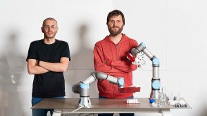 Two men stand with their arms crossed behind a table that has robot arms on